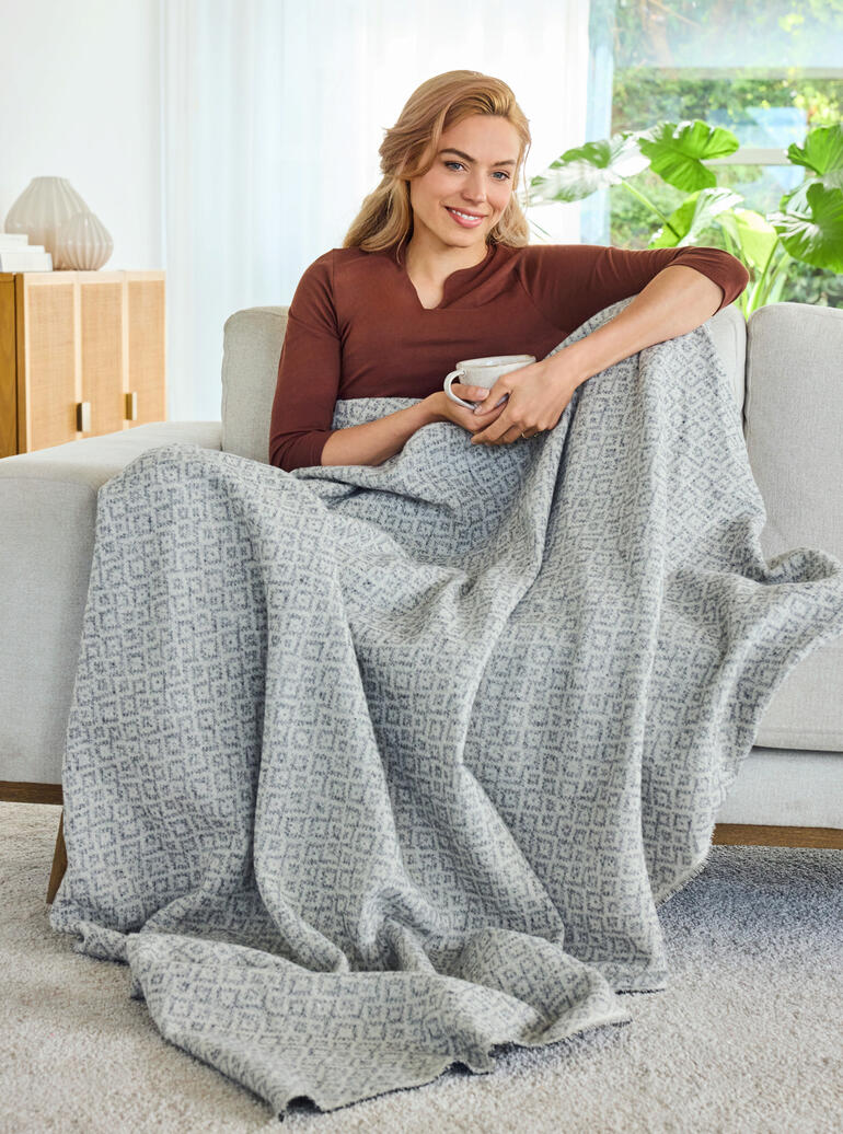 Ideal auch als leichte Decke für gemütliche Stunden auf der Couch. Frau sitzt auf einem Sofa mit einer jacquard Decke umhüllt.