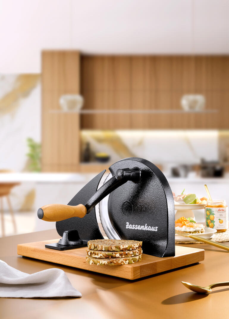 Handkurbel-Brotschneider auf einer Holzplatte mit frisch geschnittenem Brot.