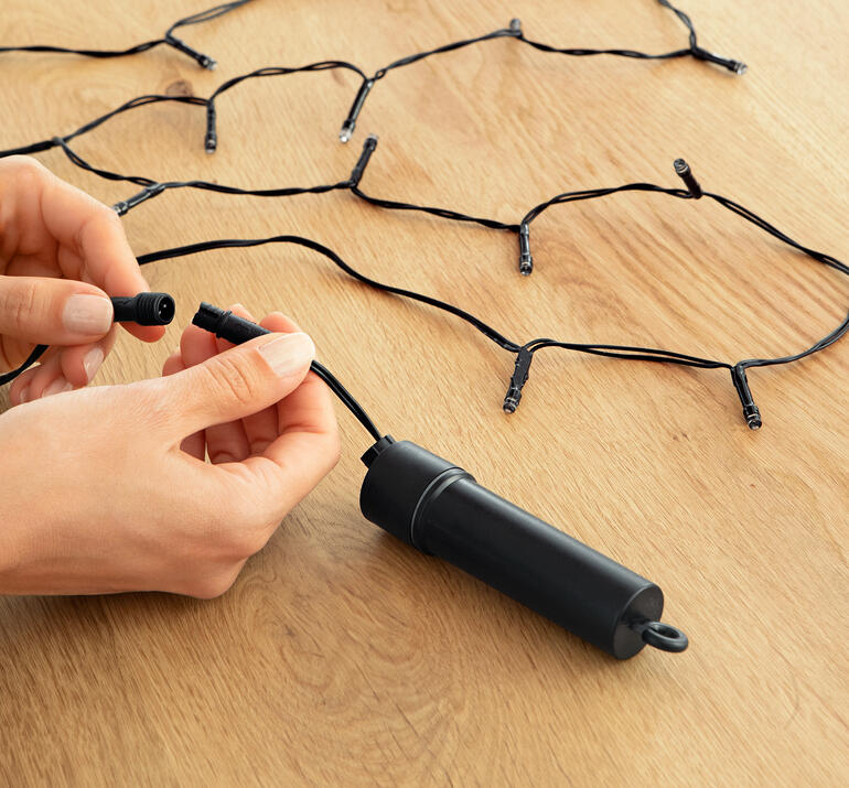Person verbindet die Lichterkette mit einem Stromkabel auf einem Holztisch.