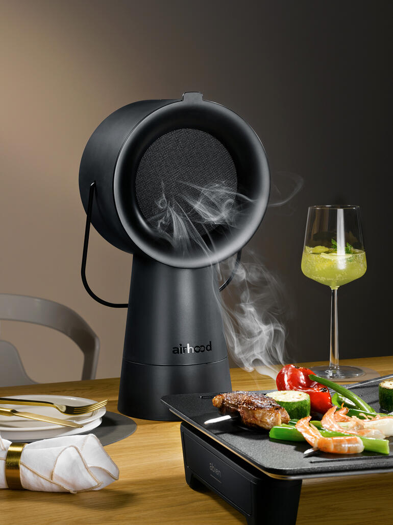 Airhood 2.0 in Schwarz, dampfendes Grillgerät auf Tisch mit Gemüse und Fleisch.