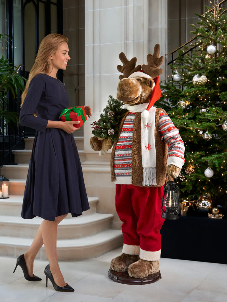Tanzender Elch in festlicher Kleidung, hält ein Geschenk und steht vor einem Weihnachtsbaum.