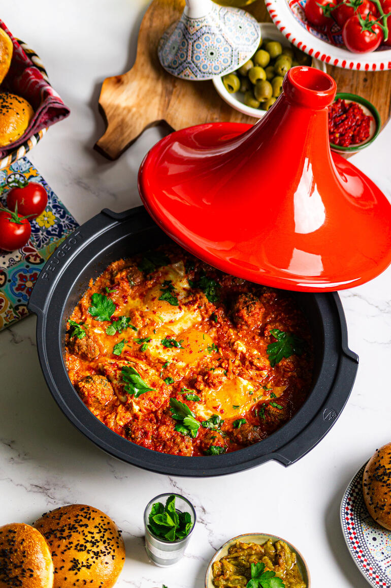 Tajine Peugeot Saveurs mit rotem Deckel, gefüllt mit buntem Gemüse und Kräutern, auf einem Tisch mit Beilagen.