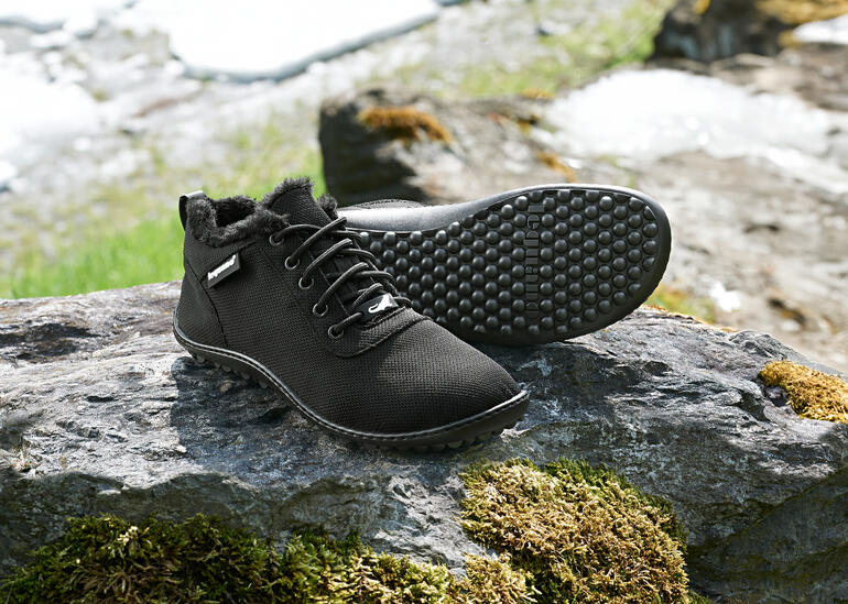 Schwarze Winter-Sneaker auf einem Stein mit Schneeresten im Hintergrund.