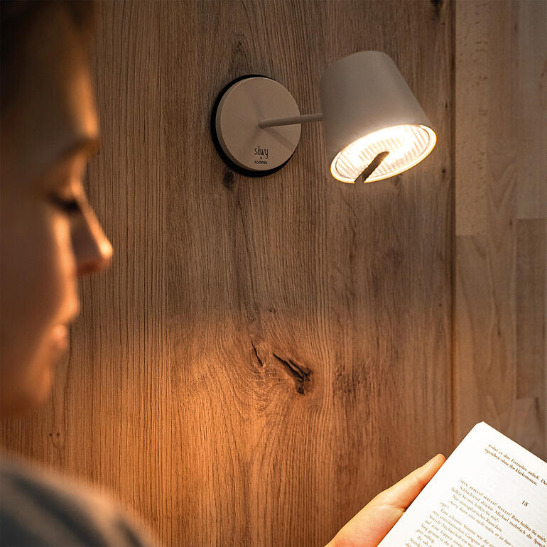 Schwarze Wandlampe mit hellem Licht über einer Leserin, die ein Buch hält.