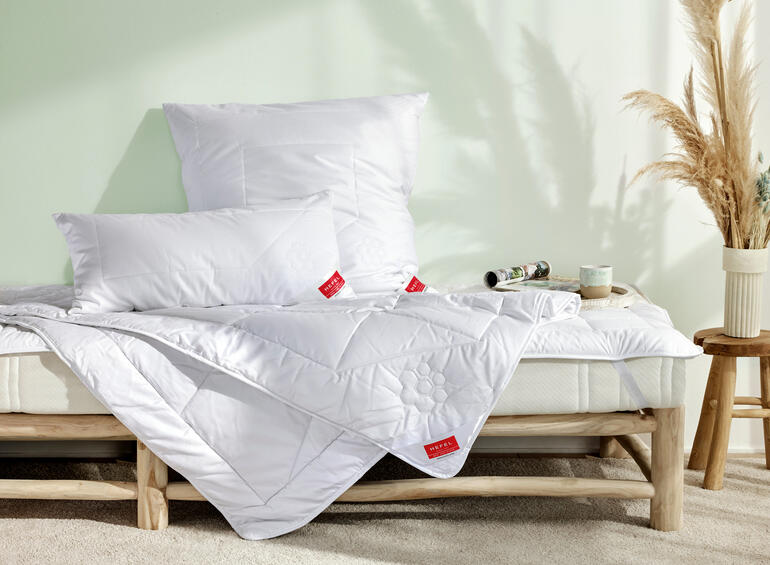 Bett mit weißen Kissen und einer Decke, helle Schlafzimmeratmosphäre.