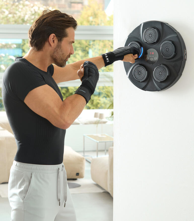 Mann schlägt mit Händen gegen Wandboxtrainer, Fitnessraum mit hellen Fenstern im Hintergrund.