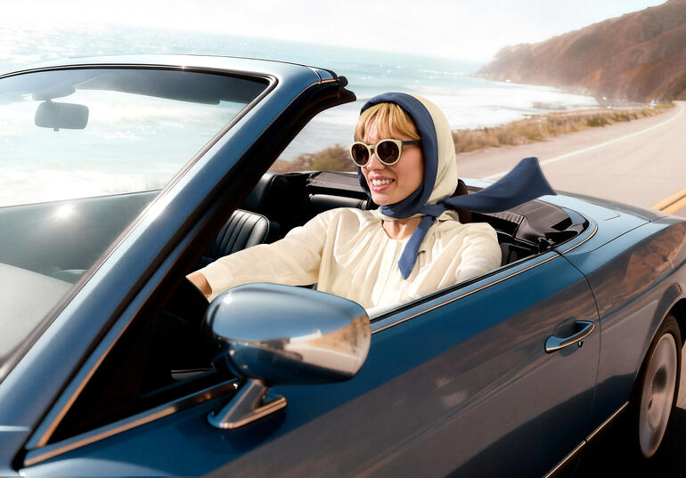 Frau in einem Cabrio mit Kopftuch und Sonnenbrille fährt an Küste entlang.
