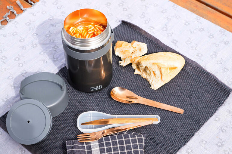 Geöffnete schwarze Snackbox mit frischer Pasta und Brot auf einem Tisch.