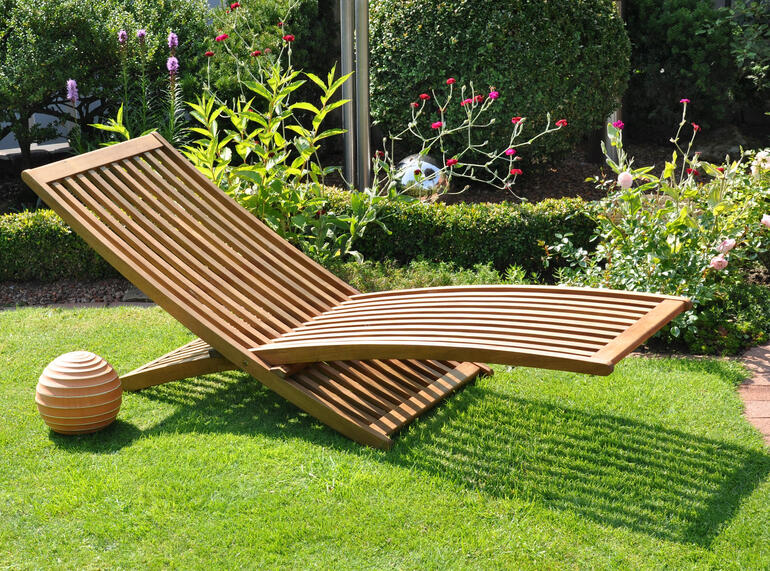 Holzsonnenliege auf grünem Rasen im Garten mit blühenden Sträuchern.