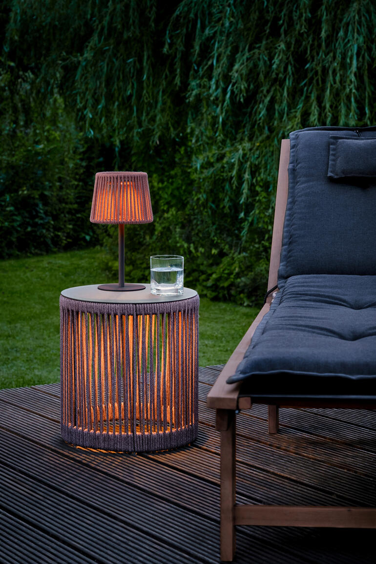 LED-Outdoor-Tisch mit orangefarbener Lampe und Wasserglas auf Holzdeck neben einer Liege.