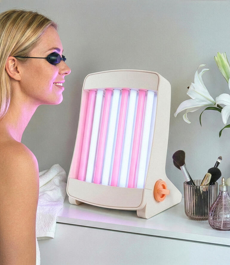 Gesichtsbräuner mit pinken und blauen Lampen, steht auf einem Tisch neben Kosmetikartikeln.