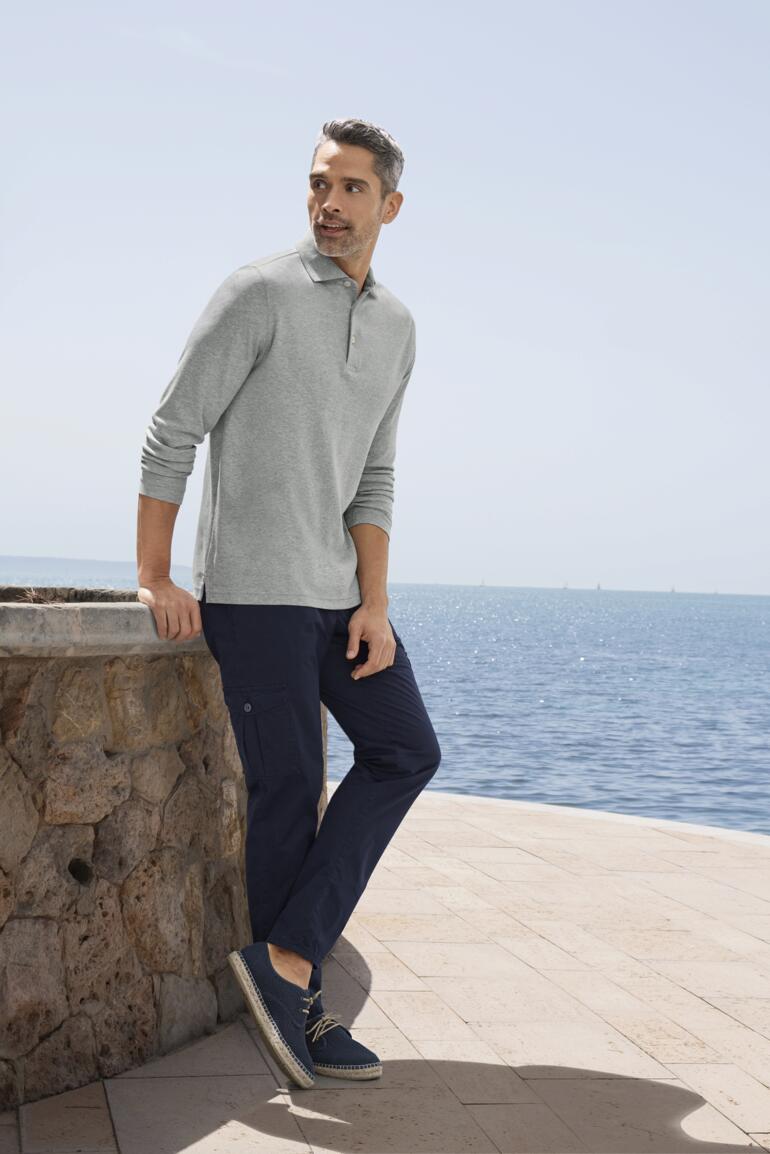 Marine Mann mit grauem Pima-Interlock-Polo, steht an einer Steinmauer mit Meerblick.