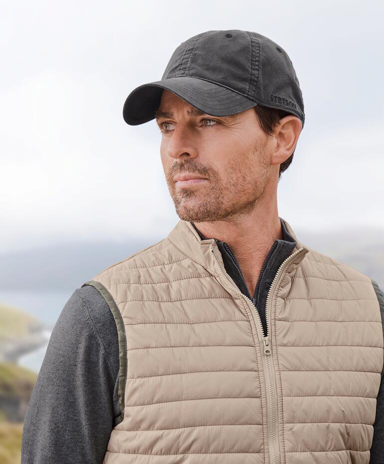 Mann mit Stetson Baseball-Cap und Weste im Freien, mit Blick auf eine Landschaft.