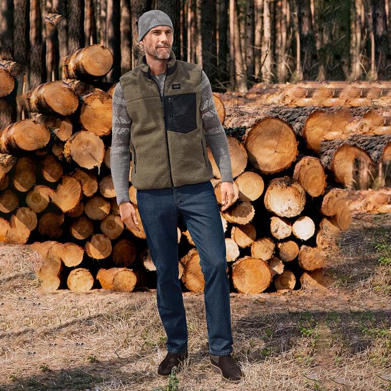 Grau Mann trägt einen Baby-Alpaka Norwegerpullover, steht vor einem Holzstapel im Wald.