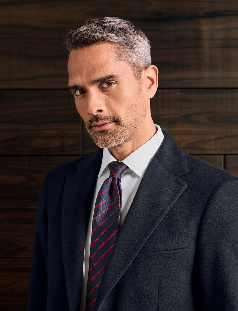 Mann mit Ascot Seidenkrawatte, skeptischer Blick, vor dunklem Holz Hintergrund.