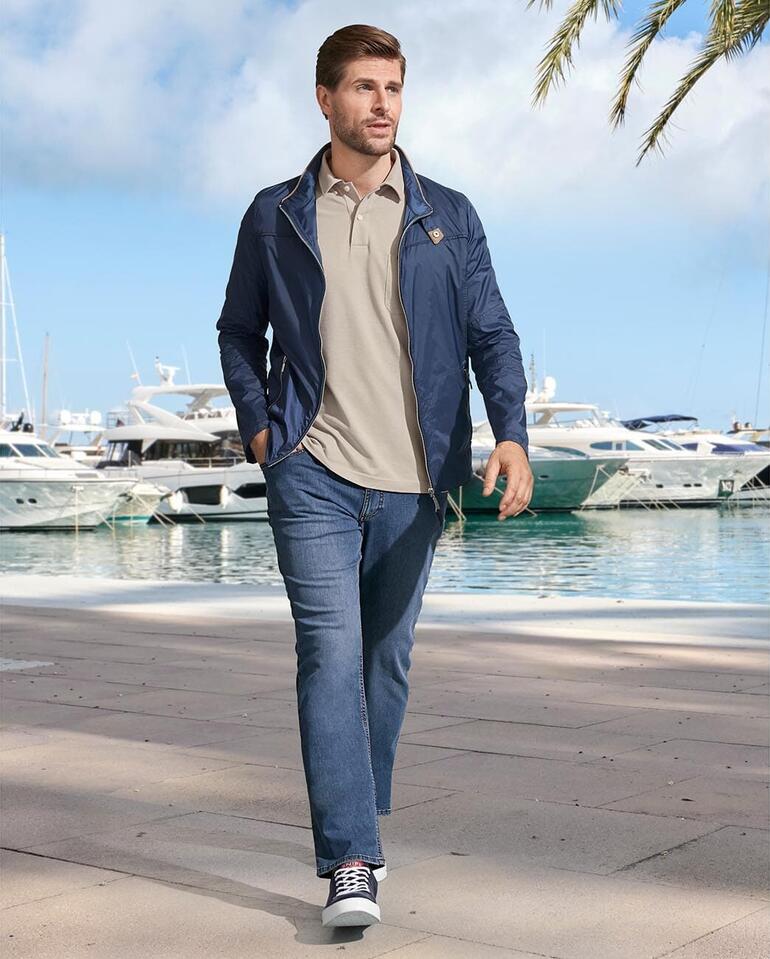 Eurex by Brax Sommer-Jeans Mann in blauer Jacke und beigem Poloshirt geht am Hafen entlang.