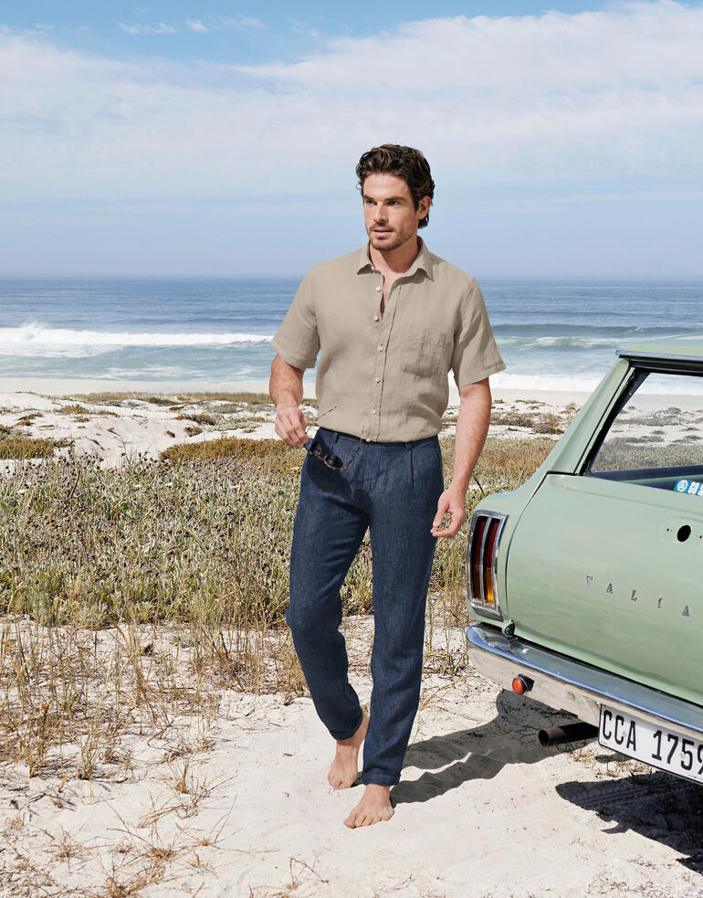 Mann mit kahler Lederjacke und Indigo-Leinenhose am Strand vor einem Auto.