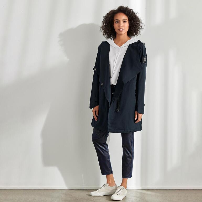 Frau im Softshell-Mantel mit Kapuze, steht in einem Raum mit hellem Licht.