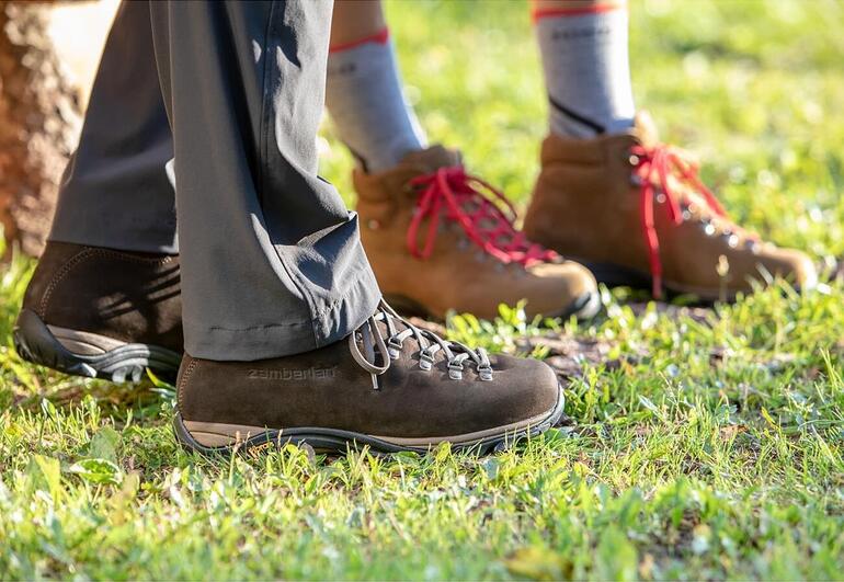 Herren, Dunkelbraun und Damen, Braun Zamberlan® Wanderstiefel, Füße einer Person in brauner und einer in beiger Stiefel auf einer Wiese.