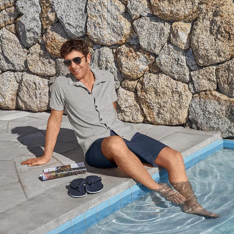 Navy Mann sitzt am Schwimmbeckenrand in einem grauen Kurzarm-Hemd und Sonnenbrille.