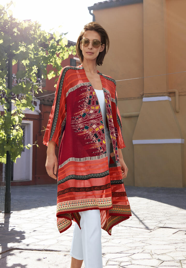 IVKO Kimono-Strickjacke Frau in Rot mit IVKO Kimono-Strickjacke, steht im Sonnenlicht auf einem Platz