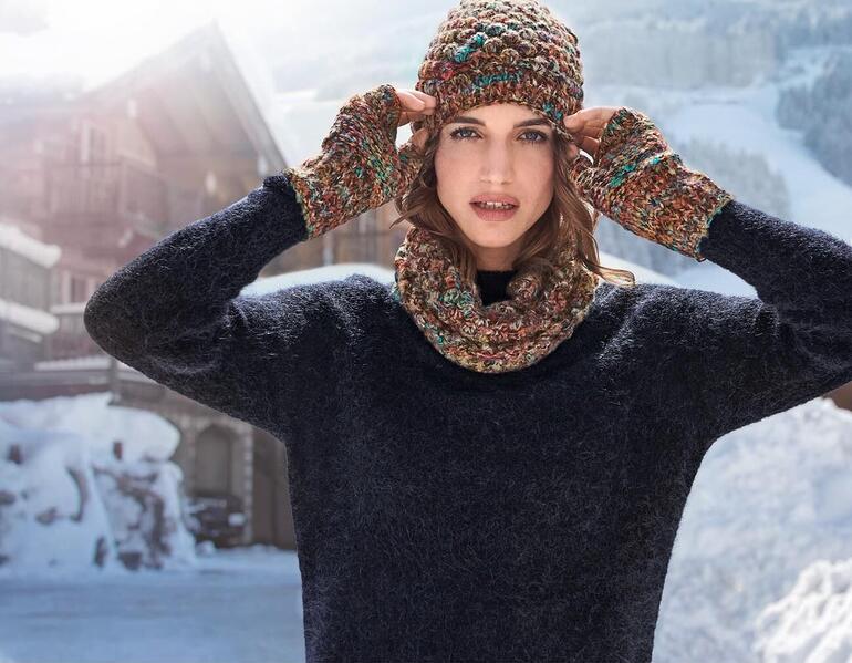 Kero Design Dornröschen-Beanie-Mütze, -Fingerlos-Handschuhe oder -Loop-Schal Frau mit bunter Mütze und Handschuhen in schneebedeckter Landschaft.