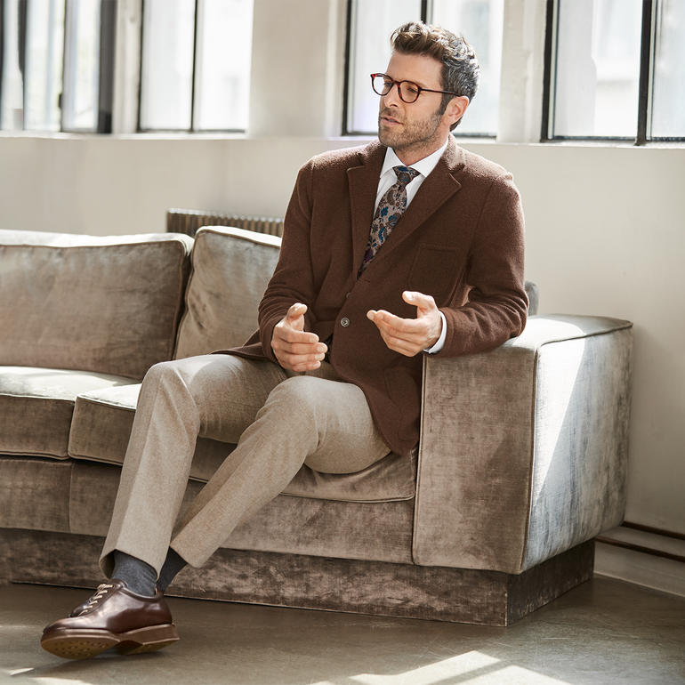 Cognac Mann trägt braunen Blazer mit einem gestreiften Krawattenmuster, sitzt auf einem Sofa.