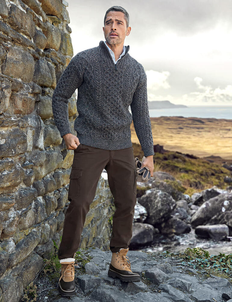IrelandsEye Aran-Troyer Mann in grauem Aran-Troyer steht vor einem Steinmauer in der Natur.