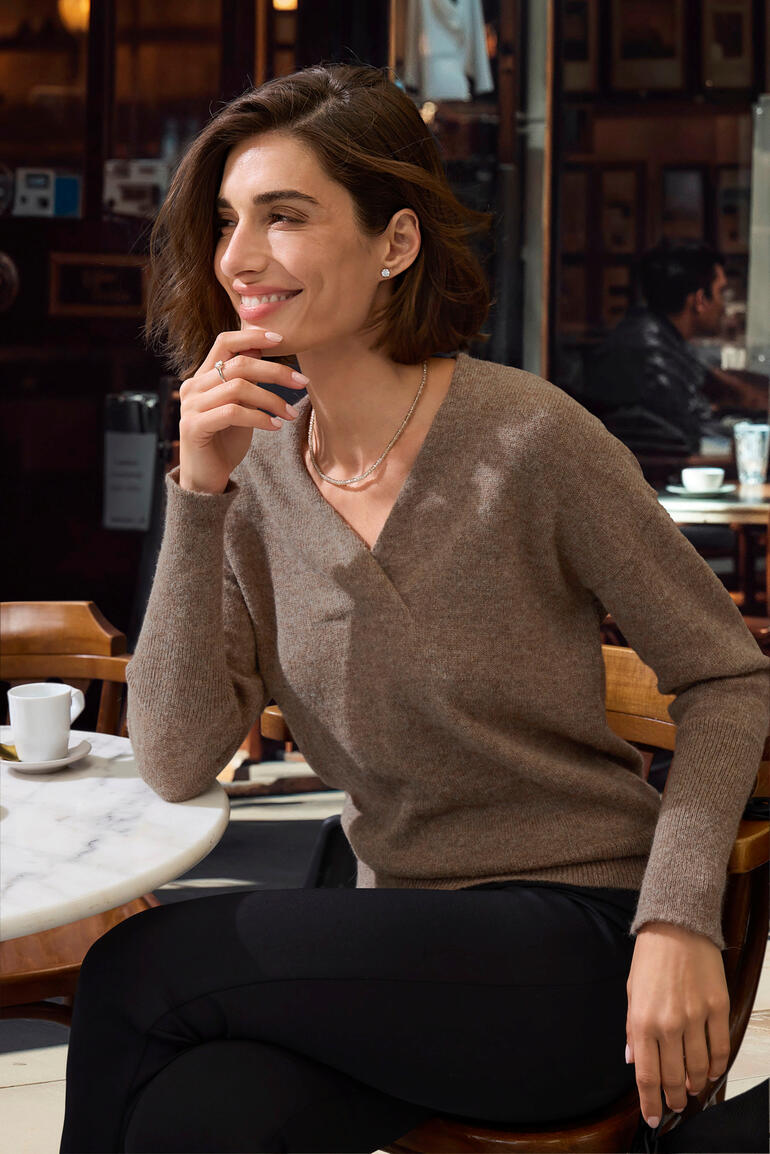 Damenpullover aus Alpaka in Braun, lächelnd am Tisch sitzend.