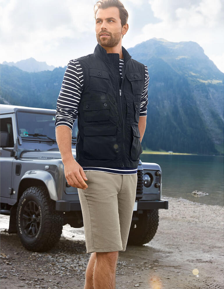 10-Taschen-Weste Mann in schwarzer 10-Taschen-Weste und gestreiftem Hemd am Bergsee.
