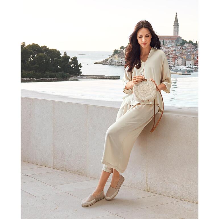 Beige Frau sitzt auf Mauer, trägt helle Kleidung und Leder-Espadrilles.