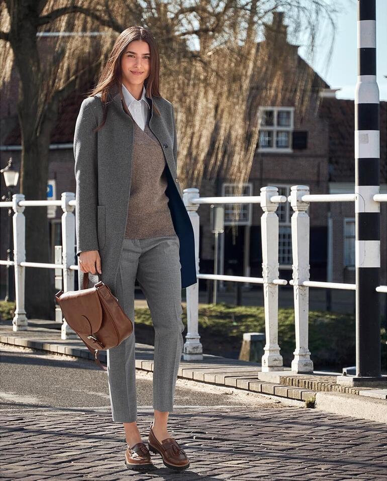 Frau in grauem Anzug mit brauner Tasche vor einer Stadtlandschaft.