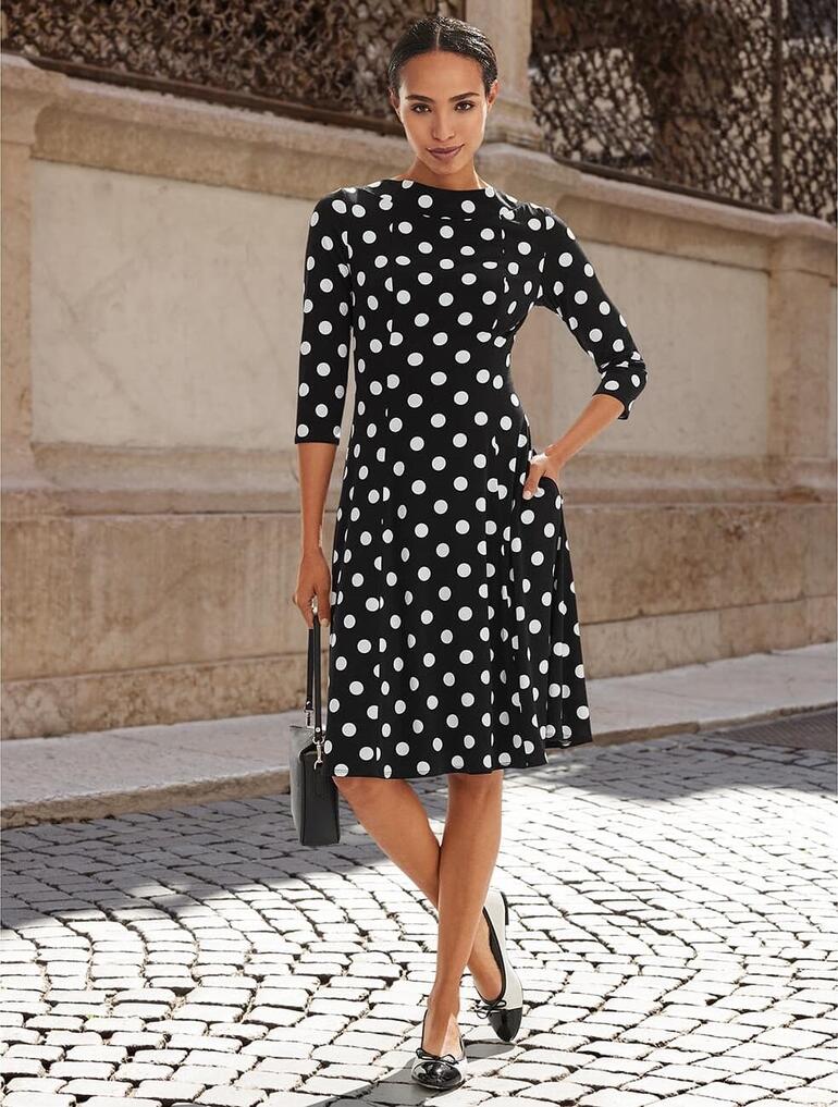 Creme/Lack-Schwarz Frau im Punktkleid steht auf einer Straße, lächelnd.