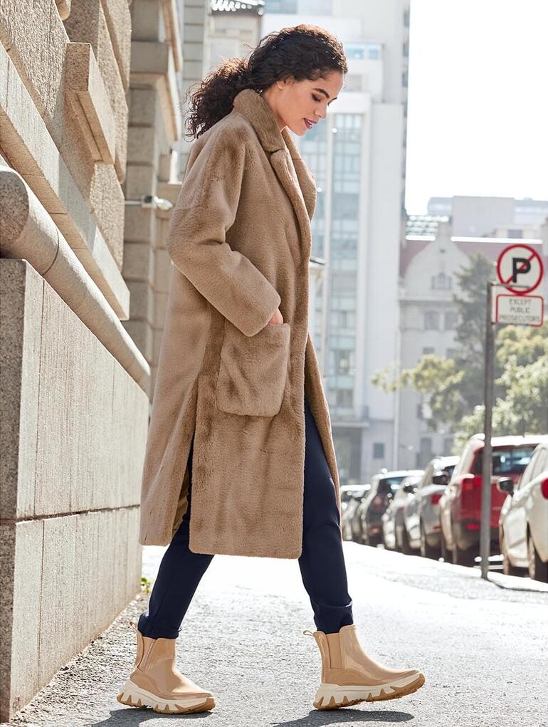 Frau in einem beigen Faux-Fur-Mantel läuft die Straße entlang