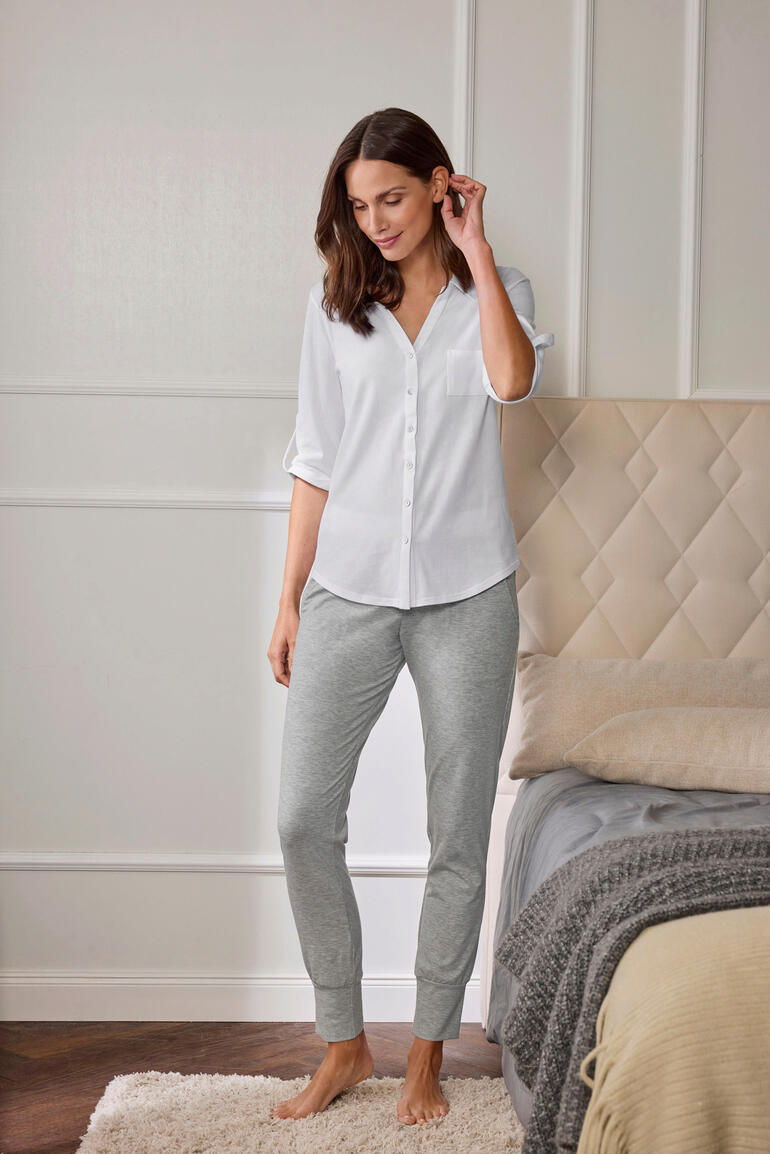 Casual-Pyjama Frau in weißem Pyjama-Oberteil und grauen Jogginghosen steht neben einem Bett.