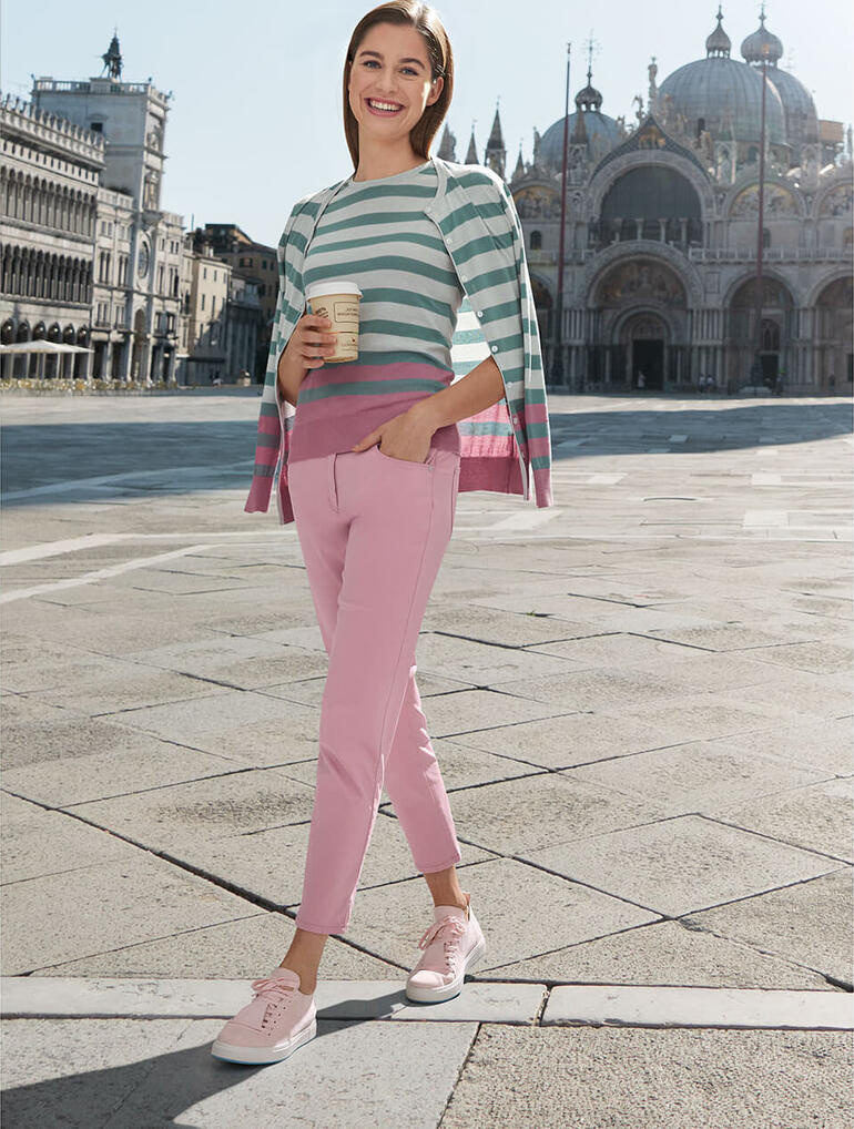Rosé Frau in einem gestreiften Oberteil und rosa Zauber-Five-Pocket-Hose steht auf einem Platz.