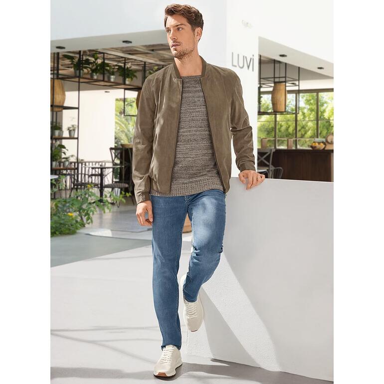 Junghans 1954 Upcycling-Pullover Mann in Amaretta-Blouson, steht an einer Wand in einem modernen Innenraum.