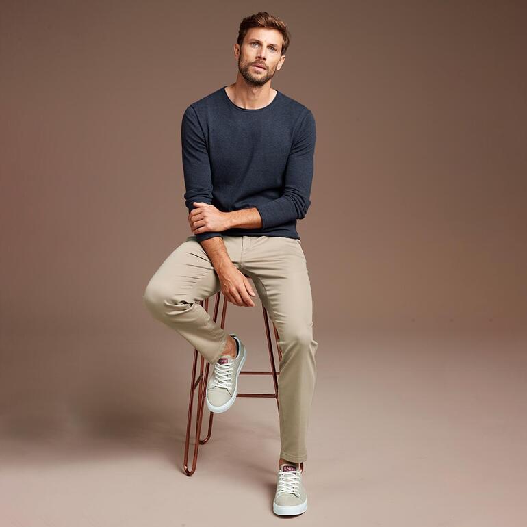 Jeansblau Mann sitzt auf Barstuhl, trägt dunkelblauen Pullover und helle Hose.
