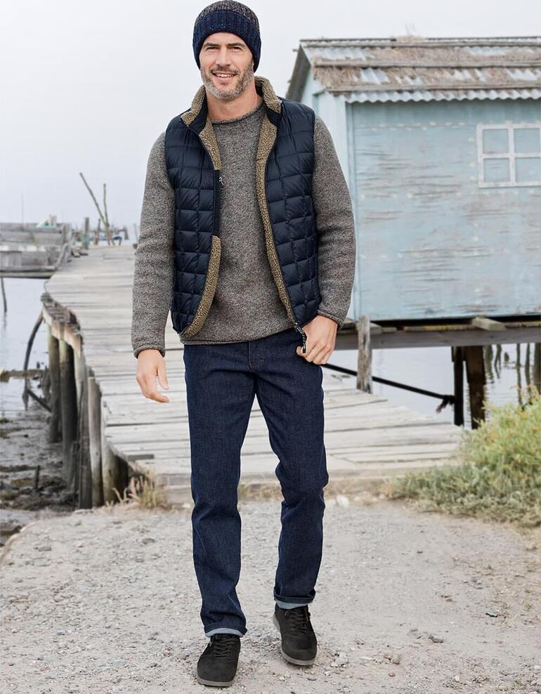 Blau Mann in Daunen-Wendeweste, grauem Pullover und Mütze, steht am Hafen.