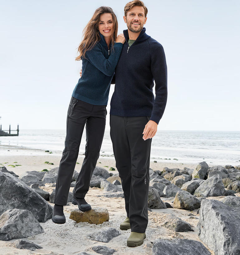 Softshell-Hose Paar in Seemannstroyern am Strand, umgeben von Steinen und Meer.