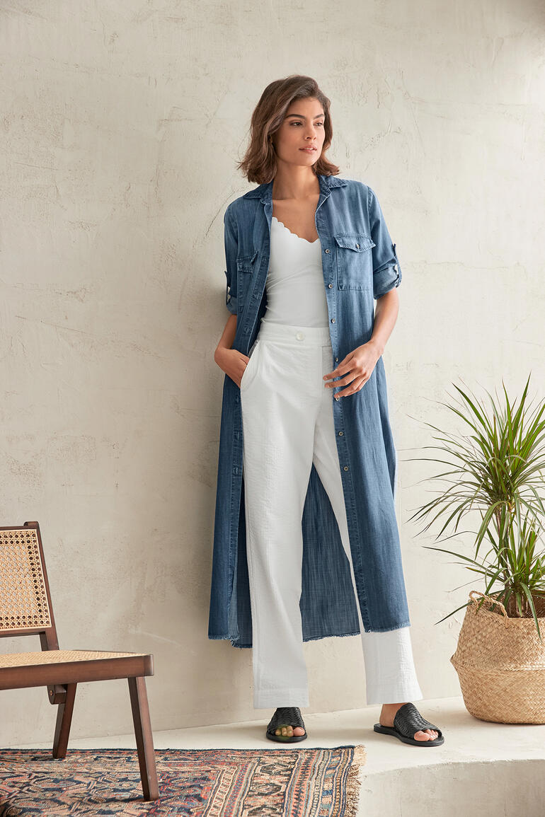 Frau im Jeansblusen-Kleid steht an einer Wand mit Pflanze.