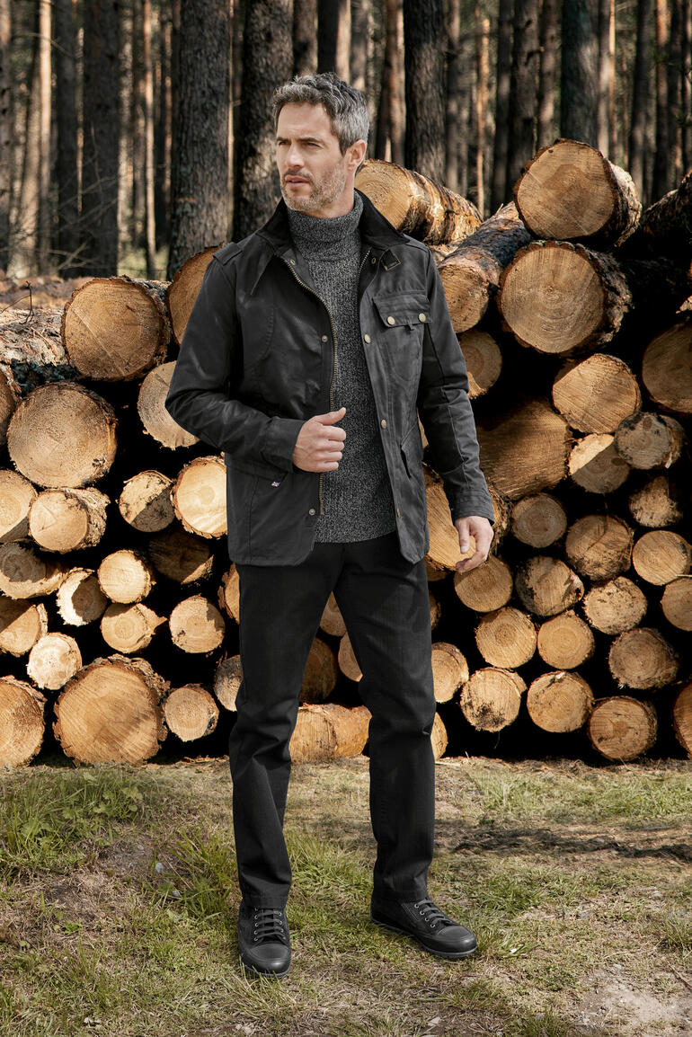 Schwarz Mann in Wachsbaum-Jacke steht in einem Wald vor Baumstämmen.