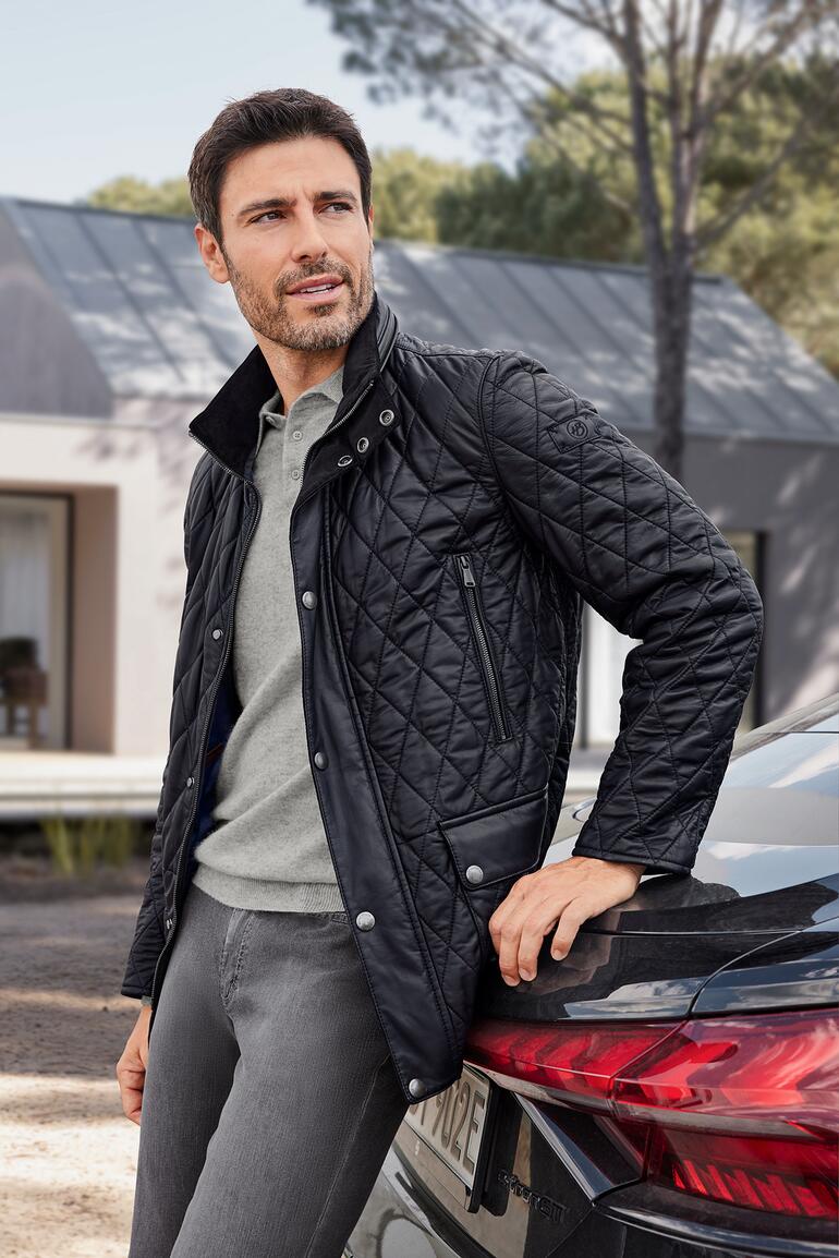 Mann in schwarzer Steppjacke mit Lammnappa vor einem modernen Gebäude.