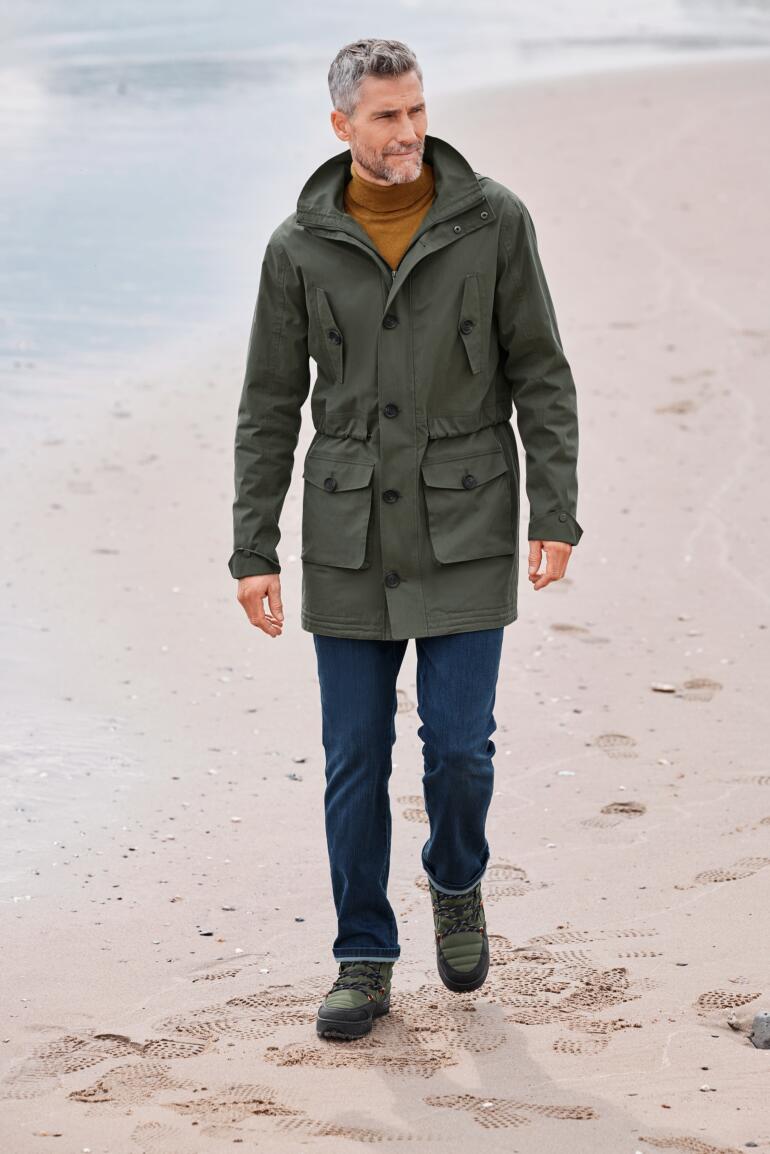 Allwetter-Baumwoll-Parka Mann in grünem Baumwollpark mit Taschen, der am Strand entlang läuft.