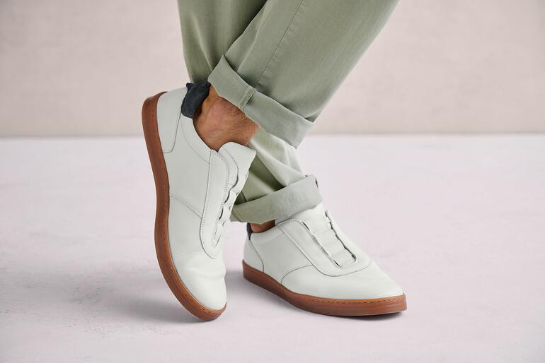 Weiß Mann trägt weiße OCONI Slip-on Schuhe auf grauem Untergrund.