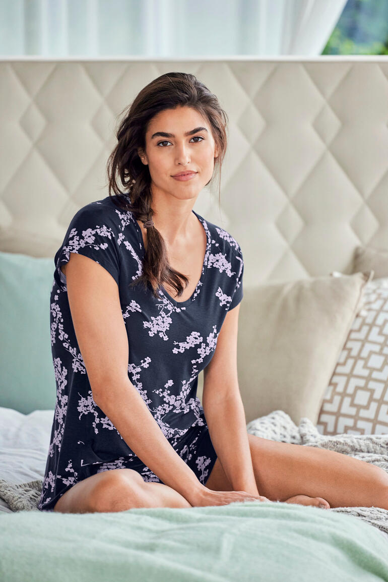 Kirschblüte Frau in schwarzem Pyjama mit Blumenmuster sitzt lächelnd auf dem Bett.