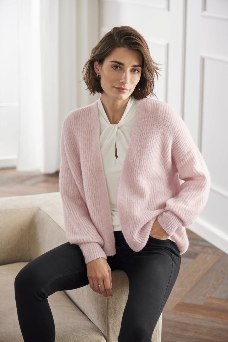 Junghans 1954 Flausch-Cardigan Frau sitzt im Junghans 1954 Flausch-Cardigan in rosa.