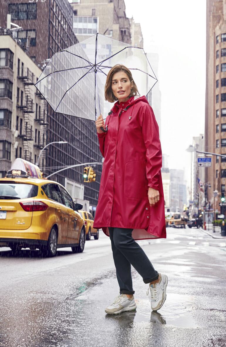 Ilse Jacobsen Red Raincoat Frau in rotem Regenmantel hält einen durchsichtigen Regenschirm in einer urbanen Umgebung.