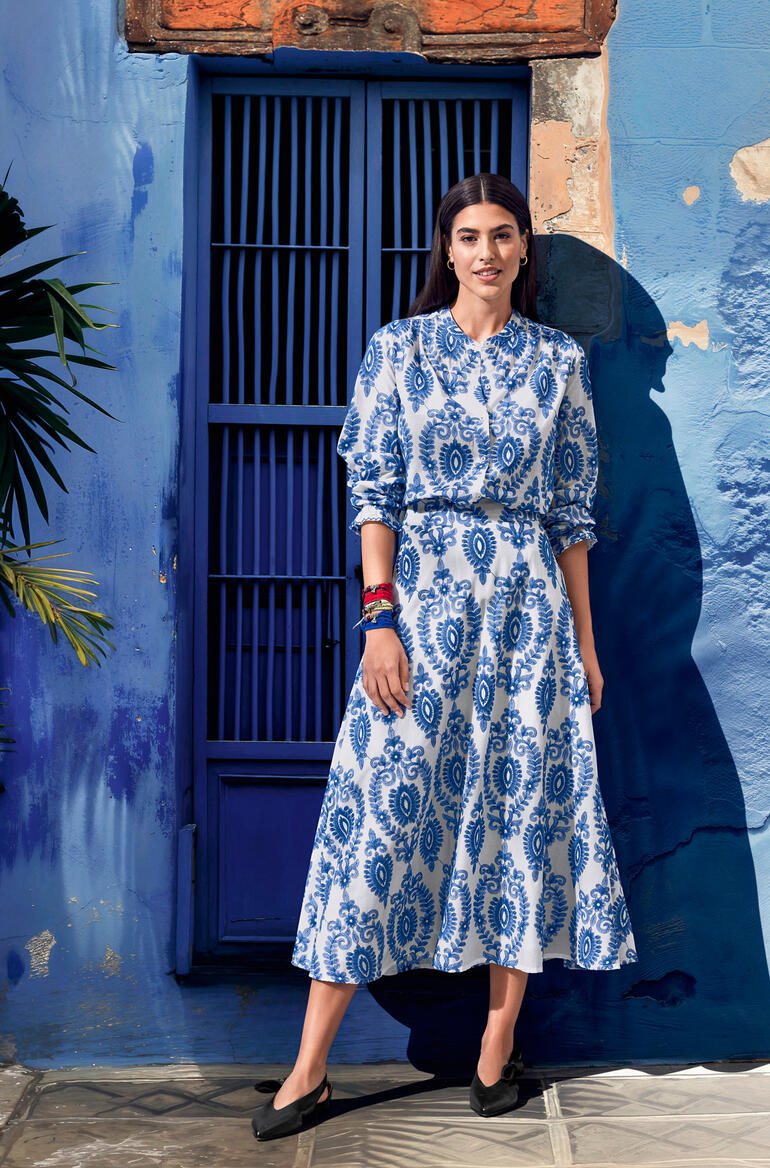 Frau in einem blauen Kleid mit weißem Muster, steht vor einer blauen Wand.