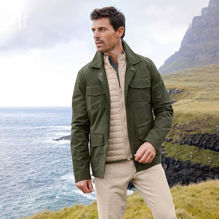 Wetterfestes Fieldjacket Mann in wetterfestem Fieldjacket steht an einer Küste mit grünem Berg im Hintergrund.